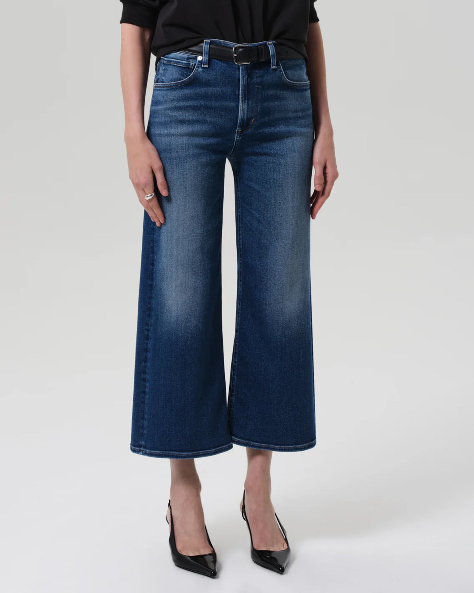 Someone wears the Lyra Wide Leg Crop in Augusta by Citizens of Humanity, paired with a black belted top and black pointed-toe heels, standing against a plain light gray background. The person's face is not visible.