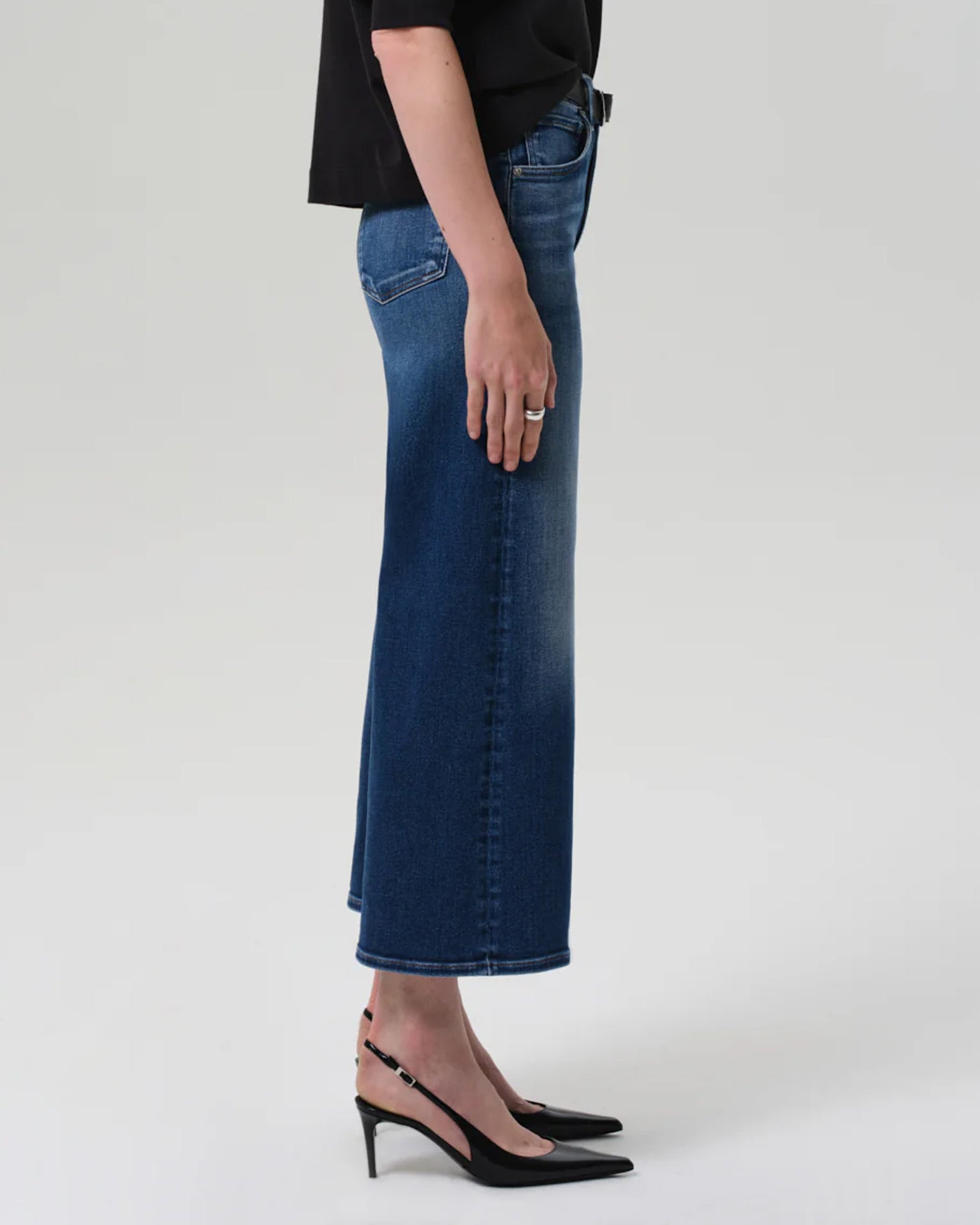 A person stands sideways against a plain light background wearing Citizens of Humanity Lyra Wide Leg Crop in Augusta jeans, a black top, and black slingback heels.