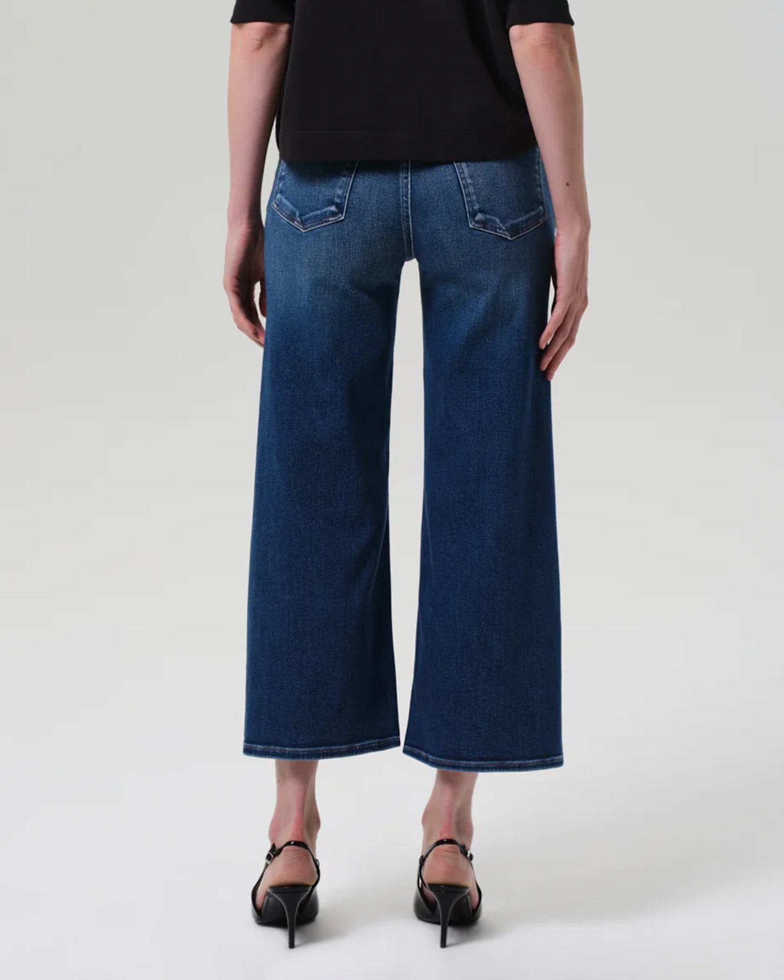A person wearing Citizens of Humanity's Lyra Wide Leg Crop in Augusta jeans and black heeled sandals, shown from the back against a plain light background.