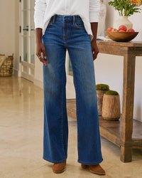 A person wears Frank & Eileen's East Galway Long Wide Leg Pant in 1999 Blue Wash with a white shirt, standing indoors on tile by a wooden table with apples and potted plants. Their head is not visible.