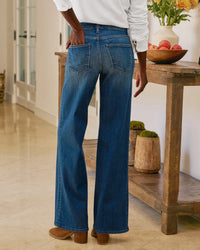 A person wears Frank & Eileen’s East Galway Long Wide Leg Pant in 1999 Blue Wash and brown heeled shoes, standing indoors on a beige floor, facing away. On the right is a rustic wooden table with vases and a bowl of apples.