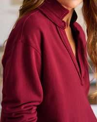 A person with long, wavy hair wears the Frank & Eileen Patrick Popover Henley in Garnet French Terry, featuring a split V-neck and raw edge collar, with a softly blurred background highlighting the upper part of the outfit.