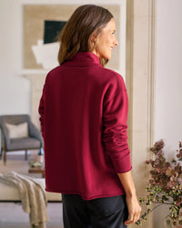 A woman with long brown hair stands indoors, facing away. She wears the Frank & Eileen Patrick Popover Henley in Garnet French Terry and dark pants. The living room background includes chairs, a couch, and a plant.