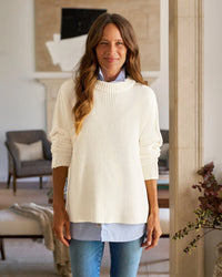 A woman with long brown hair stands indoors, smiling. She wears the Frank & Eileen Montecito Sweater in Cream over a light blue button-up shirt and blue jeans. The background reveals a living room with chairs, a couch, and wall art.