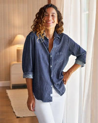 A woman with curly hair stands by a window indoors, smiling in a Frank & Eileen Eileen Relaxed Button Up Shirt in Vintage Dark Wash and white pants. One hand is on her hip; a lamp and curtains are softly lit in the background.
