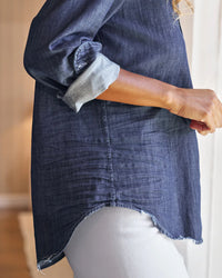 Someone stands sideways in Frank & Eileen's Eileen Relaxed Button Up Shirt in Vintage Dark Wash, featuring rolled cuffs and a frayed hem, styled with light-colored pants and their arm slightly bent.