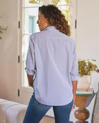 A woman with curly hair stands indoors, facing away, wearing the Frank & Eileen Eileen Relaxed Button Up Shirt in White & Navy Stripe with rolled-up sleeves and blue jeans. A table with a vase of flowers sits by a window in the background.