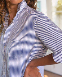 A person with curly hair wears the Frank & Eileen Eileen Relaxed Button Up Shirt in White & Navy Stripe, sleeves rolled up, standing indoors with their hand on their hip.