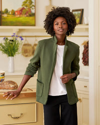 A woman with curly hair wears a Frank & Eileen Dublin Knit Blazer in Caper over a white shirt, standing in a cozy kitchen with cream cabinets. She rests a hand on a drawer beside bread, with flowers and baskets in the background.