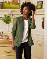 In a bright kitchen, a woman wearing a Frank & Eileen Dublin Knit Blazer in Caper and a white shirt stands touching her curly hair. Fresh bread, a mug, and flowers sit on the counter with light cabinets and shelves behind her.