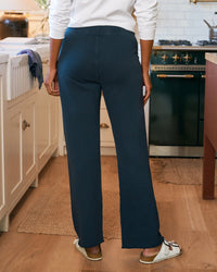 A person stands in a kitchen, viewed from behind, wearing Frank & Eileen’s Catherine Long Sweatpant in Vintage Navy with a white long-sleeve top and sandals, surrounded by white cabinets and wooden floors.