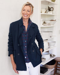 A smiling blonde woman wears a Frank & Eileen Belfast Sweatshirt Peacoat in British Royal Navy over a plaid shirt, standing in a kitchen with open shelves holding white dishes and bowls.