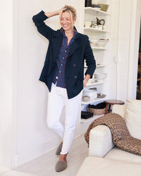 A smiling woman leans against a white wall indoors, wearing the Frank & Eileen Belfast Sweatshirt Peacoat in British Royal Navy over a plaid shirt, paired with white pants and gray slippers. Shelves and a white couch are visible in the background.
