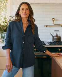 A woman with long brown hair, wearing the Frank & Eileen Eileen Relaxed Button Up in Vintage Navy Terry, stands in a bright kitchen with white tile walls, a stove, kettle, and plant in the background.