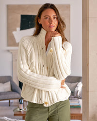 A woman with long brown hair wears the Frank & Eileen Mini Cardigan in Ivory, styled with green pants. She stands indoors, gently touching her collar and smiling, in a modern living room.