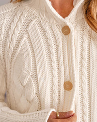 Close-up of a person wearing the Frank & Eileen Mini Cardigan in Ivory, featuring a chunky cable knit, cropped fit, and large beige buttons. The person holds the sweater near the buttons, showing part of their hand with a ring.