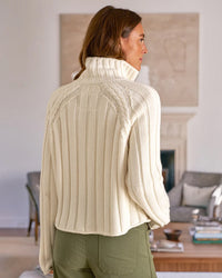 A woman with brown hair stands indoors, facing away, wearing Frank & Eileen’s Mini Cardigan in Ivory and olive green pants. The room has neutral-toned furniture and a blurred fireplace in the background.