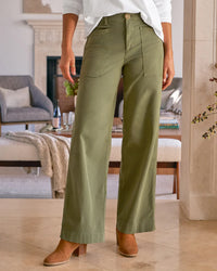 A woman stands indoors in Frank & Eileen’s Limerick Wrap Pocket Pant in Olive, paired with a white long-sleeve top and tan heels. The modern living room features sleek furniture and chic decor in the background.