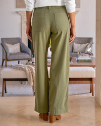 A person stands indoors facing away, wearing Frank & Eileen's Limerick Wrap Pocket Pant in Olive with light brown heeled shoes, set against a modern, neutral-toned living room with contemporary chairs and a coffee table.