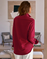 A woman with long brown hair stands indoors with her back to the camera, wearing a Frank & Eileen Eileen-Relaxed Button Up Shirt in Garnet French Terry and light pants, in a modern living room with neutral decor and seating.