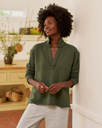 A woman with curly hair, wearing the Frank & Eileen Patrick Popover Henley in Caper Terry and light pants, stands indoors against a white wall, smiling. Behind her is a kitchen with light cabinets and potted plants.