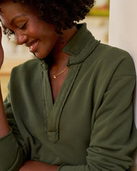 A woman with curly hair smiles gently, looking down. She wears a Frank & Eileen Patrick Popover Henley in Caper Terry and a delicate gold necklace, with a softly blurred background.