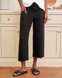 In a kitchen with light cabinets and brown tile flooring, a person wears Frank & Eileen’s Penny Patch Pocket Sweatpant in Black with a white shirt and black slide sandals, their hands resting in the sweatpant’s front pockets.