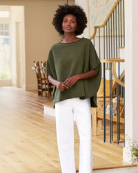 A woman with short curly hair poses confidently indoors beside a staircase, wearing Frank & Eileen's Olive Original Capelet in Caper and white pants in a modern, well-lit home with wooden floors.