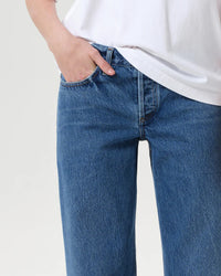 A person wearing AGOLDE’s Low Rise Loose in Charge denim blue jeans and a white t-shirt stands with one hand in their pocket against a plain light background.