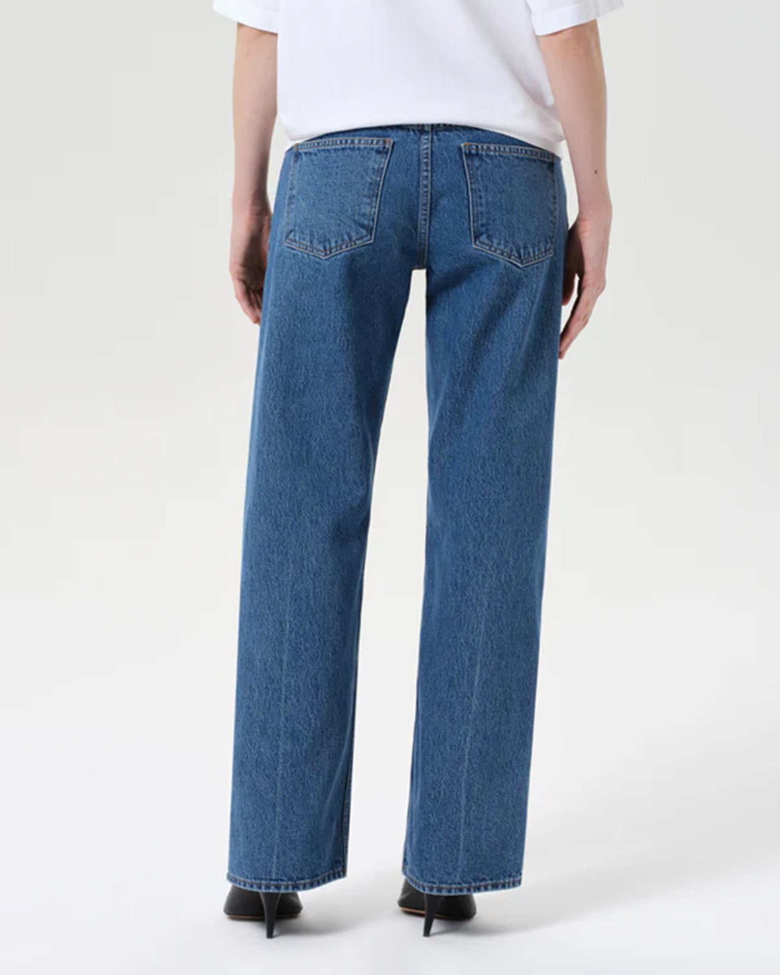 A person is seen from the back wearing AGOLDE Low Rise Loose in Charge jeans—vintage-inspired indigo, regenerative cotton denim with two back pockets—paired with a white top and black heeled shoes against a plain light background.