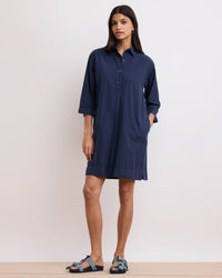 A woman stands against a plain background wearing the Hartford Rank Henley Dress in Midnight, a loose-fitting cotton poplin shirt dress with three-quarter sleeves, styled with blue sandals for a casual chic look.