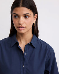 A woman with long dark hair and gold hoop earrings exudes casual chic in the Hartford Rank Henley Dress in Midnight, glancing slightly to the side against a plain light background.