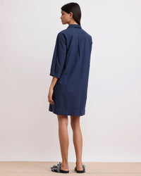 A woman stands in a plain room, facing away, wearing the Hartford Rank Henley Dress in Midnight—crafted from loose-fitting cotton poplin with three-quarter sleeves—paired with flat sandals.