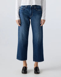 A person wearing AMO's Billie in Metaphor mid rise straight leg cropped blue jeans with black loafers and a white long-sleeve shirt, standing against a plain light background.