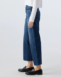 A person in AMO’s Billie in Metaphor jeans, a white long-sleeve top, and black loafers stands sideways against a plain light background.