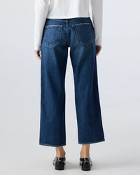 A person wearing AMO's Billie in Metaphor dark blue mid rise straight leg jeans and a white long-sleeve top is standing with their back to the camera. The jeans have large back pockets, paired with black shoes.