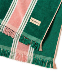 The Beach Blanket - Bistro Green Stripe by Business & Pleasure Co features green, pink, and white stripes, fringe edges, a sewn label, and is made from 100% cotton for fast drying.