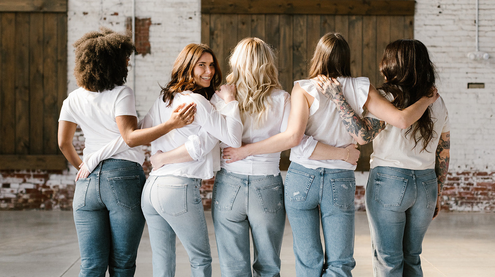 Five women in white tops and blue jeans standing together, one facing the camera while the others have their backs turned.