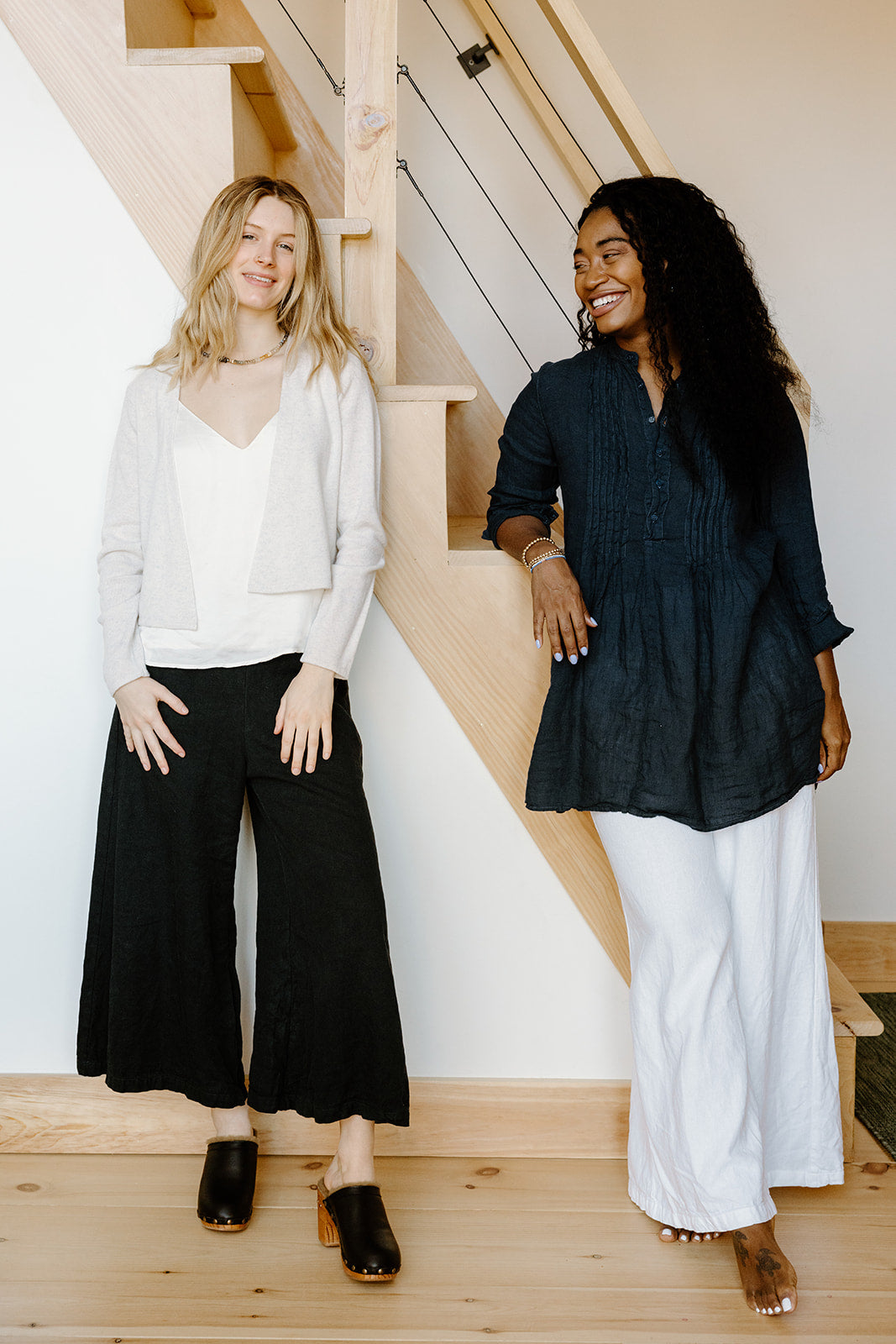 Two women smiling and standing on wooden stairs, one dressed in a light top with black pants, the other in a dark blue blouse with white pants.