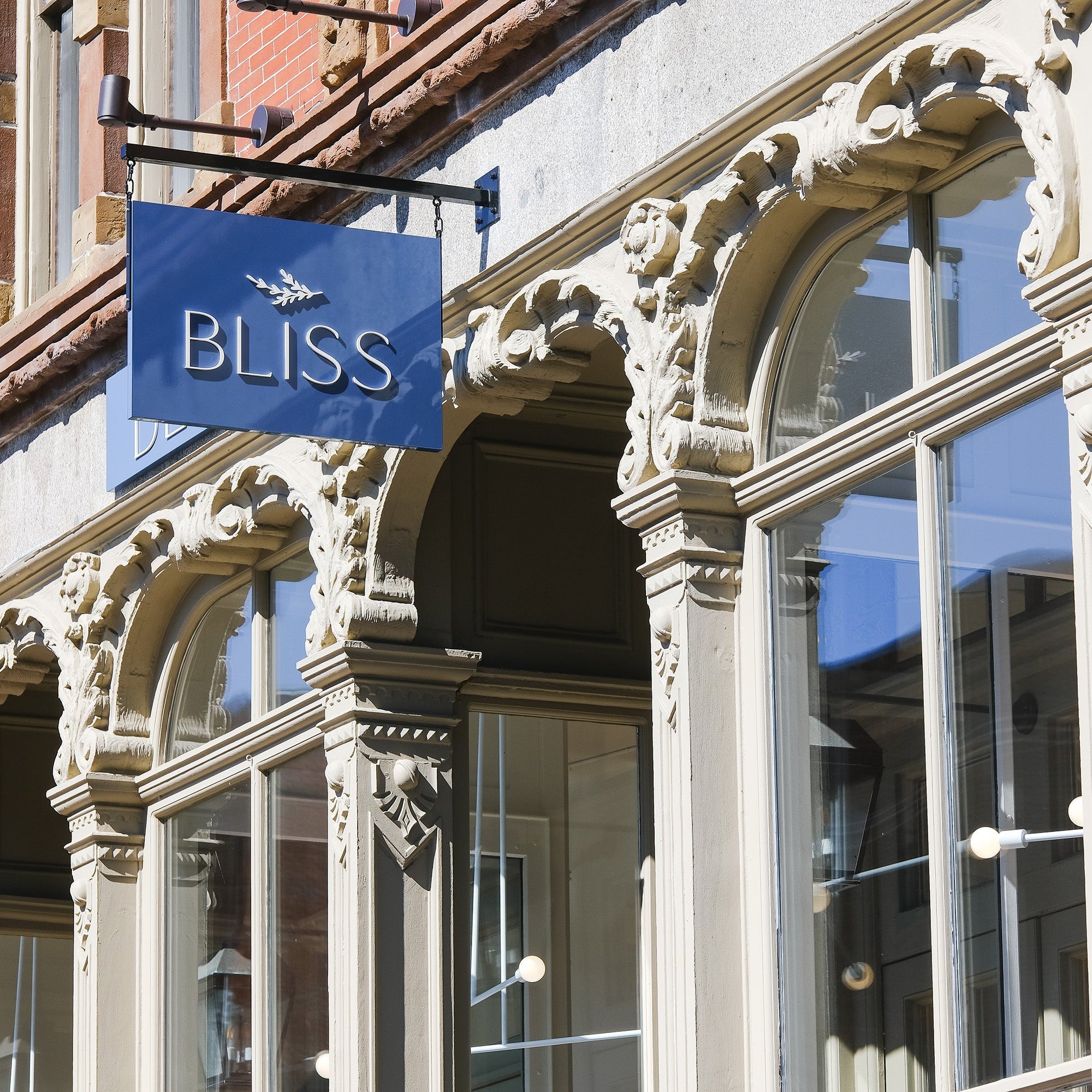 A blue sign reading BLISS with a leaf logo hangs above the ornate arched windows of a historic building in bright sunlight.