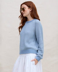 A woman with long red hair wearing sunglasses and a Margaret O'Leary Thermal Sweatshirt in Denim stands against a plain beige background, facing left with one hand in her skirt pocket.