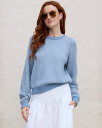 A woman with long red hair and sunglasses wears a Margaret O'Leary Thermal Sweatshirt in Denim and a white skirt, standing against a plain beige background with one hand in her pocket.