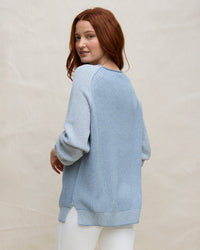 A woman with long red hair models the Margaret O'Leary Baseball Tunic in Denim, standing slightly turned to the side and looking over her shoulder against a light plain background.