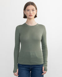 A person with short brown hair wears a fitted Margaret OLeary Shirtail Waffle Crew in Sage and blue jeans, standing against a plain white background.