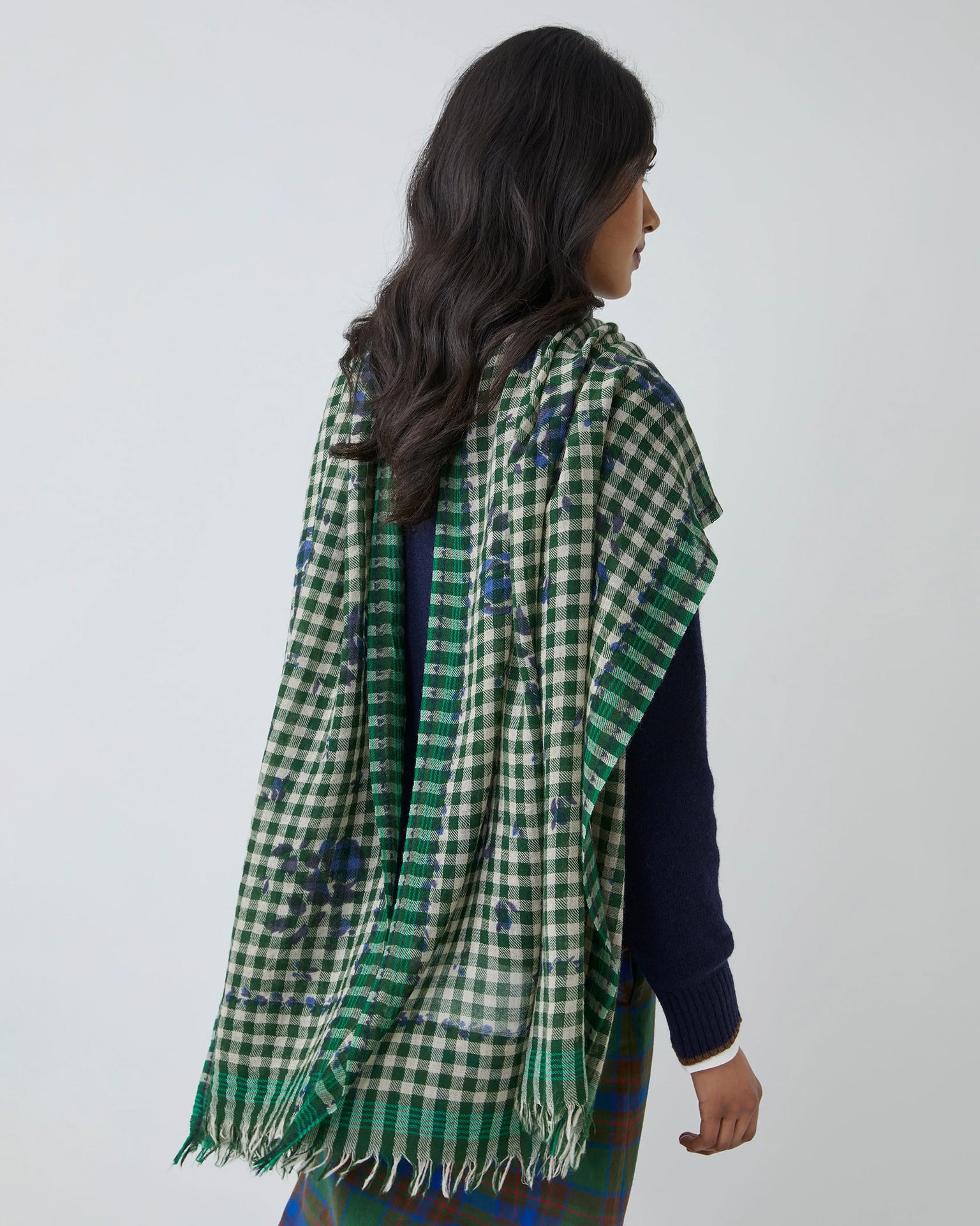A woman with long, wavy dark hair is seen from the back wearing the Moismont Vichy Wool Scarf No 693 in Flowers British Green, its fringed edges draped over her shoulders, layered atop a navy sweater and checkered skirt.