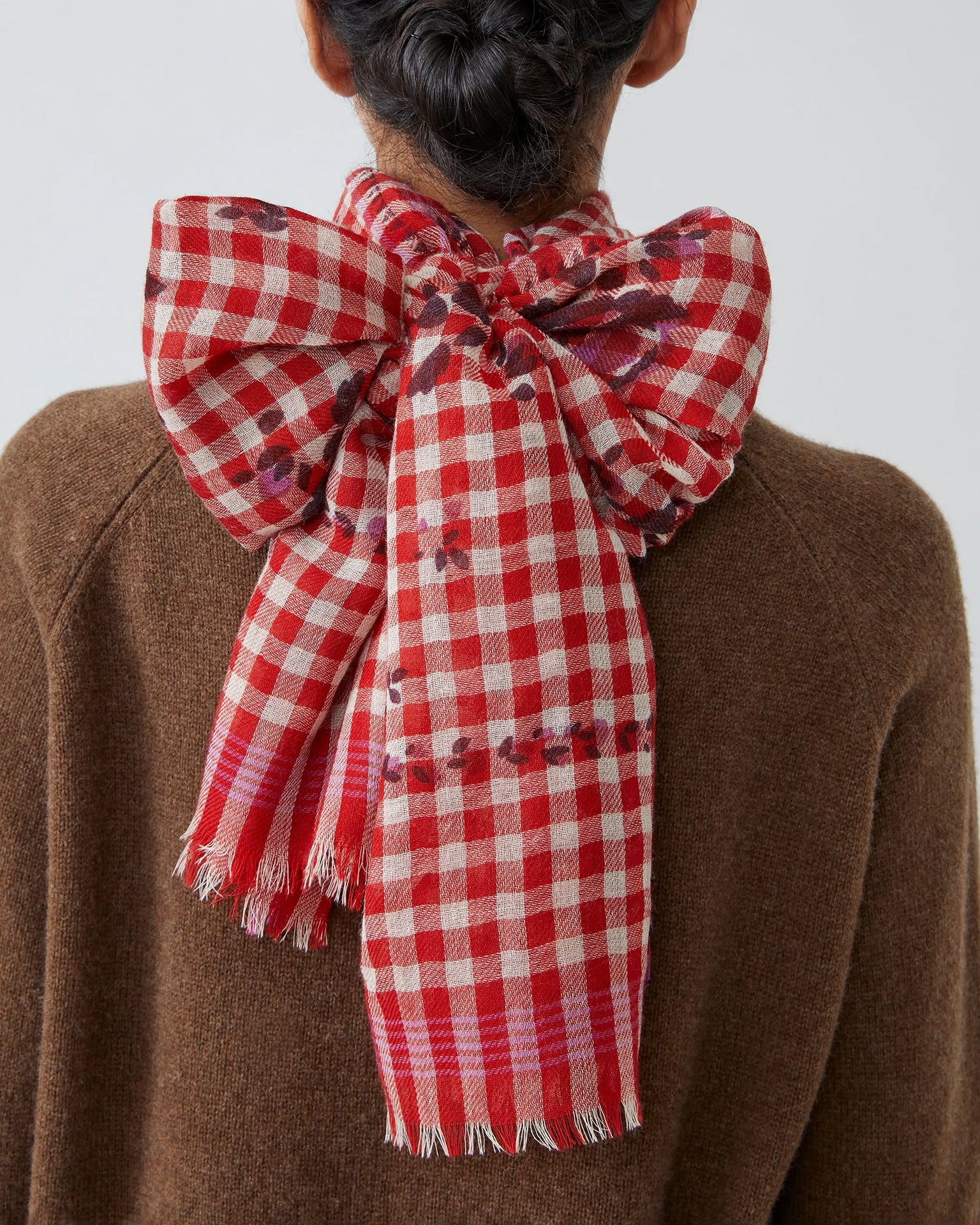 A person with dark hair in a braided updo wears a brown sweater and the Moismont Vichy Wool Scarf No 693 in Flowers Lipstick, tied in a large bow at the back. The red-and-white checkered scarf features fringed edges and subtle small patterns.