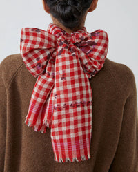 A person with dark hair in a braided updo wears a brown sweater and the Moismont Vichy Wool Scarf No 693 in Flowers Lipstick, tied in a large bow at the back. The red-and-white checkered scarf features fringed edges and subtle small patterns.