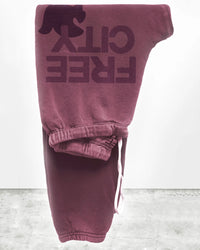 The FC Large Sweatpant in Roses by Free City features maroon French Terry fabric, a drawstring waist, and FREE CITY with a bird silhouette on the upper thigh. Folded presentation, minimalistic background. Made in USA.