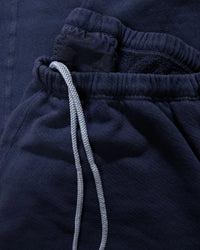 A close-up of the Basic Goodness OG Sweatpant in Moonshrooms by Free City shows navy French terry joggers with white drawstrings and a slightly open pocket revealing the waistband of another dark loungewear piece inside.