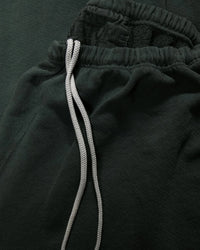 Close-up of Free City Basic Goodness OG Sweatpant in Tent, featuring a dark green French terry fabric, elastic waistband with black text, and white drawstrings laid on top.